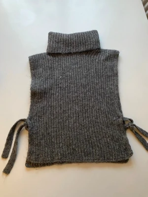 Grå stickad krage från Lindex - Mysig grå stickad kr soplugg från Lindex med polokrage och knytband i sidorna. Tillverkad i en mjuk ullmix som håller dig varm och ger en trendig lager-på-lager-look. Perfekt att dra över en hoodie eller jacka för extra värme.