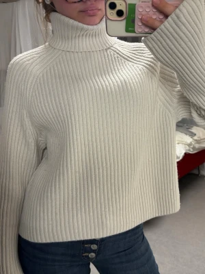 Vit ribbstickad polotröja - Säljer en krämvit ribbstickad polotröja med oversized passform och vida ärmar. Tröjan har hög polokrage och är gjord i ett mjukt, tjockt material som håller dig varm. Perfekt till jeans eller kjol för en trendig look.