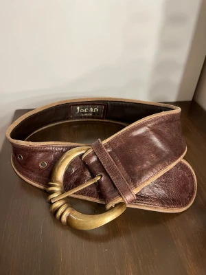 Vintage skärp i skinn från Jocasi - Snyggt brunt skärp i äkta skinn från Jocasi med bred design och stor rund mässingsfärgad metallspänne. Skärpet har flera hål för justering. 85cm😊