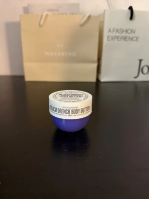 Sol de Janeiro Delicia Drench Body Butter - En liten burk Delicia Drench Body Butter från Sol de Janeiro med Cheirosa 59-doft. Burken är lila med vitt lock och har en rund form. Perfekt för dig som vill ha återfuktad hud med en lyxig känsla och tropisk doft.