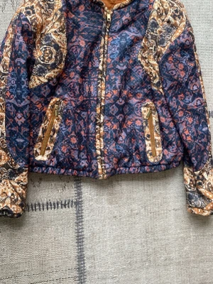 Mönstrad tunn jacka från H&M - Snygg jacka från H&M med färgstarkt mönster i blått, orange och beige. Jackan har dragkedja framtill, två fickor med guldiga dragkedjor och elastisk nederkant. Perfekt för dig som gillar unika plagg med coola detaljer.