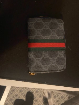 Gucci plånbok med GG-mönster - Snygg plånbok från Gucci i mörkgrått tyg med klassiskt GG-mönster. Har en grön och röd rand på framsidan och guldfärgad dragkedja. Perfekt storlek för kort och kontanter, och ger en lyxig känsla till din vardag.