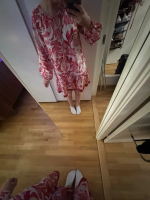 Rosa och vit blommig klänning - Säljer en luftig kortklänning med rosa och vitt blommönster. Klänningen har långa ärmar med resår vid handlederna, v-ringning med knytband och volangdetalj nedtill. Perfekt för dig som gillar färg och mönster!