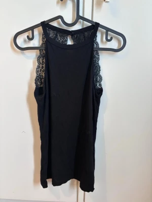 Svart spetsdetaljerad linnetopp - Snygg svart linnetopp med spetsdetaljer längs axelbanden och övre delen av ryggen. Toppen har hög halsringning och är ärmlös, vilket ger en trendig och elegant look. Perfekt att matcha med jeans eller kjol för en cool outfit.