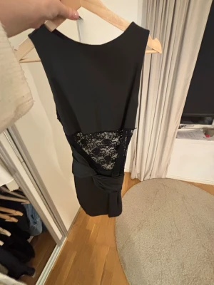 Svart kort klänning med spetsdetalj - Snygg svart kort klänning med spetsdetalj i midjan och omlottkänsla nedtill. Ärmlös modell med rund halsringning. Perfekt för dig som vill ha en elegant och stilren look.