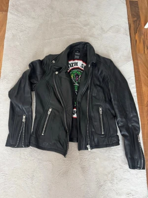 South Side Serpents skinnjacka - Snygg svart skinnjacka inspirerad av South Side Serpents från Riverdale. Jackan har flera silvriga dragkedjor, klassisk bikerkrage och ett stort broderat märke på ryggen med texten 'South Side Serpents' och en grön orm. Perfekt för dig som vill sticka ut med en edgy look.