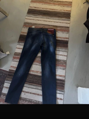 Mörkblå jeans från Dsquared2 - Säljer ett par snygga mörkblå jeans från Dsquared2 med klassisk femficksmodell och diskreta slitningar. Jeansen har en rak passform och detaljer i kontrastsömmar samt en stor läderpatch med logga bak i midjan.