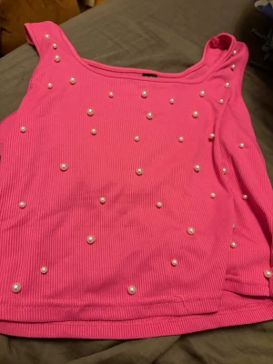Rosa ribbad pärlprydd linnetopp - Snygg rosa linnetopp med ribbad struktur och dekorativa pärlor över hela framsidan. Toppen är ärmlös och har en croppad passform som passar perfekt till höga jeans eller kjol. Perfekt för dig som vill sticka ut lite extra.