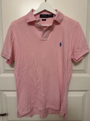 Pikétröja från Ralph Lauren - Klassisk rosa pikétröja från Ralph Lauren med blå broderad logga på bröstet. Slim fit-modell i mjuk bomull som passar perfekt till jeans eller shorts.