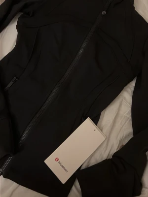 Svart zip-jacka från Lululemon - Snygg svart träningsjacka från Lululemon. Jackan har figurnära passform, långa ärmar och praktiska sidofickor med dragkedja. Helt ny, köpt i butik med prislappen kvar. I storlek 2. 