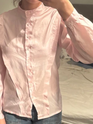 Ljusrosa blus med puffärm - Söt ljusrosa blus med puffiga långärmar och ståkrage. Använd 2 gånger. Blusen har knappar hela vägen framtill och är gjord i ett tunt, glansigt material som ger en elegant känsla. Perfekt att styla med jeans eller kjol för en trendig look.