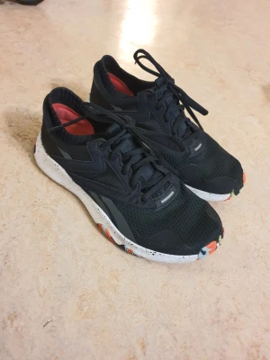 Svarta Reebok sneakers med färgglad sula - Säljer ett par svarta Reebok sneakers med färgglad yttersula i orange, svart och grönt. Skorna har lågt skaft och röda detaljer på insidan. Perfekta för dig som vill sticka ut med coola detaljer.