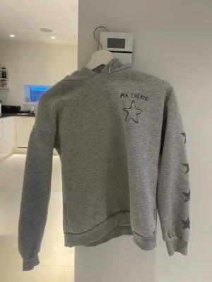 Grå hoodie med stjärntryck - (Tröjan går inte längre att köpa)Snygg grå hoodie med huva och trycket 'MA CHÉRIE' samt en svart stjärna på bröstet. På ena ärmen finns flera stjärntryck i svart. Tillverkad i mjukt sweatshirtmaterial och har ribbade muddar vid ärmslut och nederkant. Perfekt för en avslappnad och trendig look.OBS ⚠️: lite trasig vid halsen!!! 