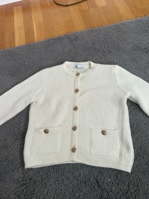 Krämvit kofta från Zara Knit - Stilren krämvit kofta från Zara Knit med rund halsringning och träknappar framtill. Koftan har två fickor med dekorativa knappar och är stickad i ett mjukt material. Perfekt att slänga över en t-shirt för en clean look.
