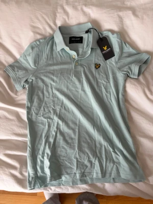Ljusblå pikétröja från Lyle & Scott - Snygg ljusblå/turkos pikétröja från Lyle & Scott med klassisk krage, korta ärmar och knappslå framtill. Tröjan har den ikoniska gula loggan broderad på bröstet och är tillverkad i mjuk bomull. Perfekt till jeans eller shorts. Tveka inte på att höra av dig vid funderingar!