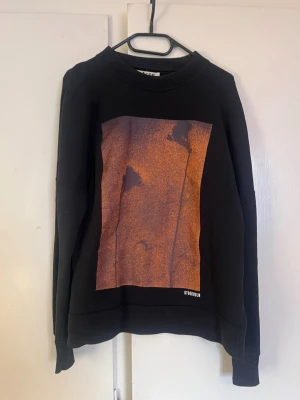 Svart sweatshirt Acne Studios - Cool svart sweatshirt från Acne Studios med en stor grafisk print i orange och brunt framtill. Tröjan har rund halsringning och ribbade muddar vid ärmslut och nederkant. Perfekt för dig som gillar streetwear och stilrena detaljer.