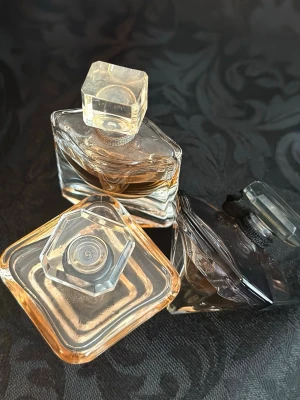 Lancome parfymflaskor i glas 3st (5 ml varje) - Tre eleganta parfymflaskor från Lancome i transparent glas med geometriska former och dekorativa lock. Flaskorna har olika former, bland annat fyrkantig och diamantformad, och innehåller parfymer i bärnstensfärgade toner. Perfekt för dig som gillar snygga flaskor och lyxiga dofter.