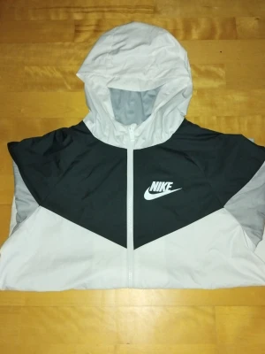 Nike vindjacka med huva svart/grå - Snygg vindjacka från Nike i svart och ljusgrått med vit dragkedja och huva. Jackan har en sportig design med Nike-logga på bröstet, elastiska muddar och två fickor med dragkedja. Perfekt för blåsiga dagar och streetwear-stil. Jackan har tecken på användning i form av små fläckar längst ner bredvid dragkedjan, se bild sju. Hör av er vid några frågor kring jackan!