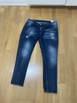 Mörkblå Dsquared2 jeans med färgstänk 1:1 - Säljer ett par mörkblå jeans från Dsquared2 med coola färgstänk och slitningar på både fram- och baksida. Jeansen har smal passform, klassiska fem fickor och är tillverkade i denim. Perfekta för dig som vill ha en unik och trendig look.