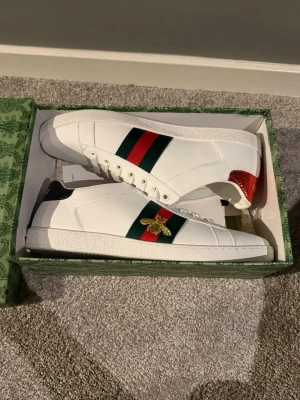 Gucci Ace vita sneakers - Säljer ett par vita Gucci Ace sneakers i läder med klassiska gröna och röda ränder på sidan och broderad guldfärgat bi. Baksidan har metallicröd och metallicgrön hälflik med ormskinn. riktigt sjysst inför hösten/vintern! Bara att skriva om ni har frågor eller vill ha fler bilder!