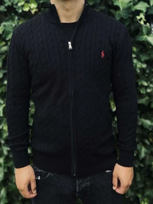 Oanvänd Svart kabelstickad tröja Polo Ralph Lauren - Kabelstickad svart tröja från Polo Ralph Lauren med hel dragkedja framtill och röd broderad logga på bröstet. Tröjan har lång ärm, ribbade muddar och en smal passform. Perfekt för dig som gillar klassisk och stilren design.