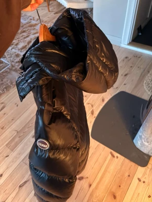 Svart dunjacka från Colmar - Snygg svart dunjacka från Colmar med hög krage och stor huva. Jackan har en quiltad design med breda kanaler och dragkedja framtill. Perfekt för kalla vinterdagar och riktigt skön att bära. Klassisk och stilren modell som passar till det mesta. Luvan är avtagbar. Storleken är 48 men sitter som en S/M