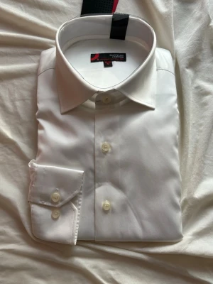 Vit skjorta i egyptisk bomull Dressmann - Klassisk vit skjorta från Dressmann i mjuk egyptisk bomull. Skjortan har normal passform, lång ärm och knappar framtill samt vid manschetten. Perfekt till skjorta och slips eller mer avslappnat med uppkavlade ärmar.