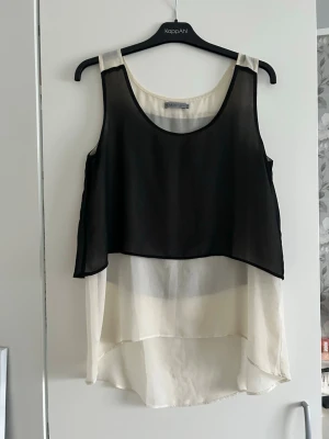Svartvit ärmlös linnetopp i lager - Snygg ärmlös linnetopp i två lager med svart transparent tyg över vitt. Toppen har rund halsringning och längre bakstycke för en cool look. Perfekt att styla med jeans eller kjol för en trendig outfit.