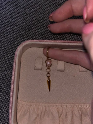 Guldörhänge med rosa pärla och spike - Unikt örhänge i guldfärgad metall med en kedja, rosa pärldetalj och en spetsig spike längst ner. Snyggt och edgy smycke som sticker ut och passar perfekt för dig som gillar att mixa stilar. Örhänget har en enkel ring som fäste.