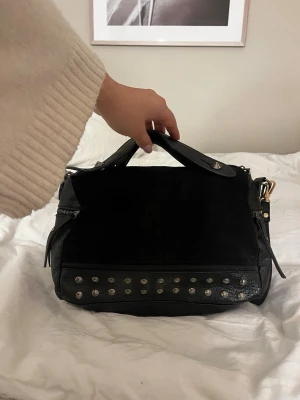 Svart handväska med nitar - Cool svart handväska i mocka och läder med silverfärgade nitar längs nederkanten. Väskan har dubbla handtag och dragkedjedetaljer på sidorna. Perfekt för dig som gillar en edgy stil och vill ha plats för allt du behöver.
