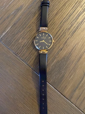 Svart klocka från Mockberg i läder - Stilren klocka från Mockberg med svart urtavla och guldfärgade detaljer. Armbandet är i äkta läder och har en klassisk design med justerbar längd. Perfekt accessoar för dig som gillar minimalistisk och elegant stil.