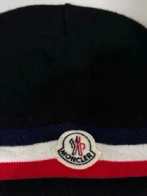 Svart Moncler mössa med logga - Säljer en svart Moncler mössa med klassisk Moncler-logga framtill. Mössan har en ribbad kant med blå, vit och röd rand som ger en sportig och stilren look. Perfekt accessoar för dig som gillar exklusiva märken och vill hålla dig varm.