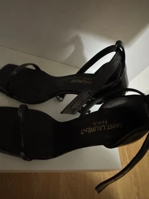 Svarta Saint Laurent sandaler - Stilrena svarta sandaler från Saint Laurent med smala remmar och fyrkantig tå. Klack och justerbart ankelband ger en elegant look. Tillverkade i blankt läder som ger en lyxig känsla. Perfekta för dig som gillar minimalistisk design. Storlek 36. Använda endast två gånger på grund av att jag har knäskada och kan ej använda klackar. :(