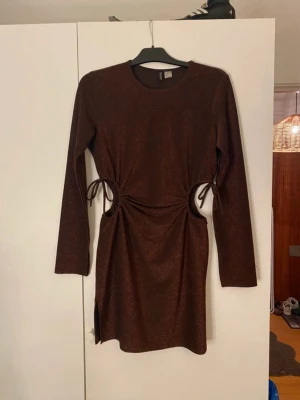 Vinröd glittrig klänning - Säljer en vinröd glittrig klänning från H&M med långa ärmar och snygga cut-outs i både midjan samt snörningsdetaljer. Klänningen har en tight passform och en liten slits på sidan samt i ärmarna. Perfekt för dig som vill sticka ut lite extra! 