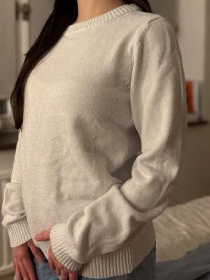 Vit stickad tröja med rund hals - Klassisk vit stickad tröja med rund halsringning och ribbade muddar vid ärmslut och nederkant. Tröjan har en avslappnad passform och är perfekt för lager på lager. Materialet ser mjukt och bekvämt ut, vilket gör den enkel att styla till jeans eller kjol.