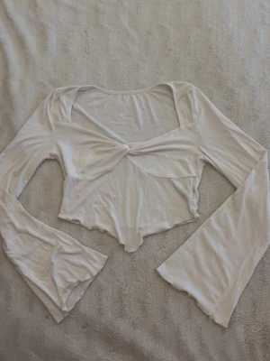 Vit croppad topp med knyt framtill - vit croppad topp med lång ärm och snygg knyt-detalj framtill. Toppen har en lätt utsvängd ärm och är tillverkad i ett mjukt, stretchigt material som känns skönt mot huden.