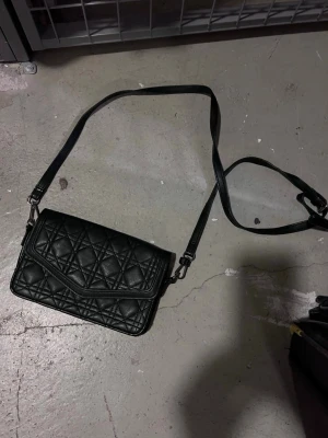 Svart quiltad axelväska - Snygg svart axelväska i quiltat mönster med justerbar axelrem. Väskan har en rektangulär form och stängs med ett lock. Perfekt för dig som vill ha en stilren och trendig accessoar till din outfit.