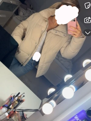 Beige pufferjacka med hög krage - Snygg beige pufferjacka med hög krage och dragkedja framtill. Jackan är väldigt varm och passar perfekt till de kalla vinterdagarna. Det finns en liten svart fläck men det går bort i tvätten eller om man gnuggar det lite. 