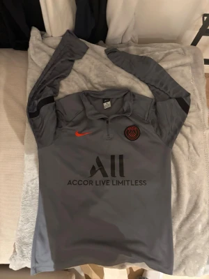 Grå långärmad PSG träningströja Nike - Snygg grå långärmad träningströja från PSG med svart och röd logga, halv dragkedja och tryck på bröstet och ryggen. Tillverkad i funktionsmaterial som passar perfekt till fotbollsträning eller gym. Diskreta svarta detaljer på ärmarna och tryckta sponsorloggor.