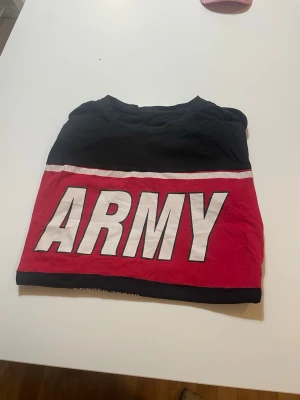 Svart och röd t-shirt med ARMY-tryck - Cool t-shirt i svart och rött med stort vitt ARMY-tryck över bröstet. T-shirten har en klassisk rund halsringning och är tillverkad i mjukt bomullstyg. Perfekt för dig som gillar streetwear och statement-plagg.