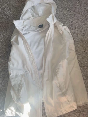 Vit vindjacka med huva från Nike - Snygg vit vindjacka från Nike i storlek XS. Jackan har huva, dragkedja framtill och två fickor. Tillverkad i lätt och tunt material med meshfoder för extra komfort. Perfekt för blåsiga dagar och enkel att matcha med olika outfits.