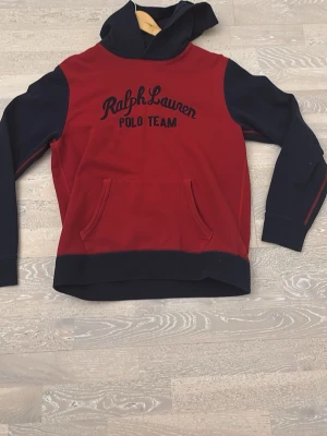 Röd och marinblå hoodie Ralph Lauren - Snygg hoodie från Ralph Lauren Polo Team i rött och marinblått. Stor huva, magficka och broderad logga framtill. Perfekt för en sportig och avslappnad stil. Mjukt material och klassisk passform.