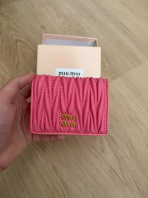 Rosa plånbok från Miu Miu - Snygg rosa plånbok från Miu Miu i quiltat läder med guldfärgad logga framtill. Plånboken har flera fack för kort och ett myntfack med tryckknapp. Perfekt storlek för att få plats i mindre väskor och riktigt trendig design.