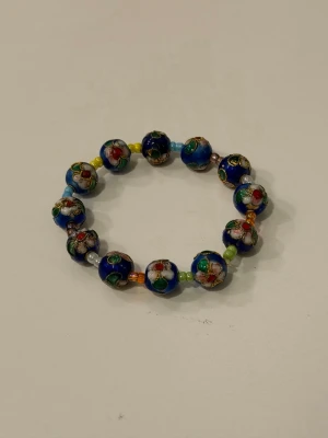Blått armband med cloisonnépärlor - Färgglatt armband med blå cloisonnépärlor dekorerade med blommönster i rött, grönt och vitt. Mellan pärlorna sitter små pärlor i olika färger. 