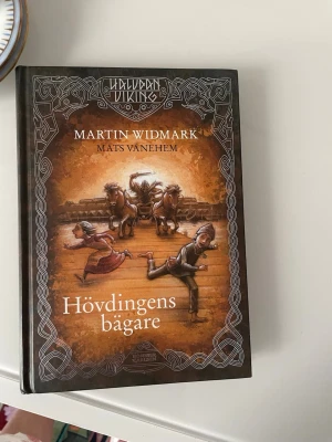 Hövdingens bägare av Widmark - Halvdans Viking Hövdingens bägare är en äventyrsbok skriven av Martin Widmark och illustrerad av Mats Vänehem. Boken har hårda bruna pärmar med vikingainspirerade illustrationer och mönster. Perfekt för dig som gillar spännande historier om vikingar och tävlingar.