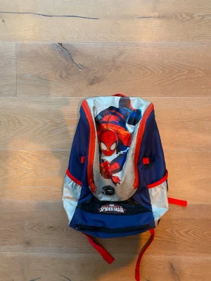 Spiderman ryggsäck i blått och rött - Cool ryggsäck med Spiderman-motiv i blått, rött och silver. Framsidan har en stor bild på Spiderman och logga, medan baksidan har ett spindelmotiv. Justerbara axelband och flera fack. Perfekt för skolan eller utflykten!
