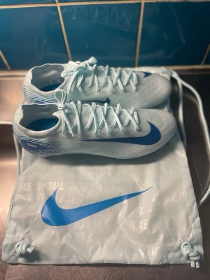 Nike Mercurial vita/blå fotbollsskor - Snygga Nike Mercurial fotbollsskor i vitt med blå detaljer och stor blå Swoosh på sidan. Skorna har en strömlinjeformad design, tunn ovandel i syntetmaterial och klassisk snörning. Kommer med matchande vit/blå Nike-påse. Perfekta för snabba spelare på planen.