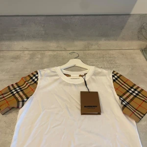 Burberry t-shirt  - Snygg vit t-shirt från Burberry med klassiska rutiga ärmar. T-shirten är i storlek M men passar S. Perfekt för dig som vill uppgradera din garderob. Tveka inte på att höra av er om ni har några funderingar. Pris kan diskuteras!