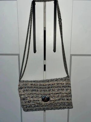 Beige tweed handväska med kedjerem - Trendig handväska i beige och blått tweedtyg med silverfärgad kedjerem. Väskan har ett klassiskt kuvertformat lock och snygg vridknapp i metall. Perfekt för dig som gillar en chic och stilren look med en touch av vintage.