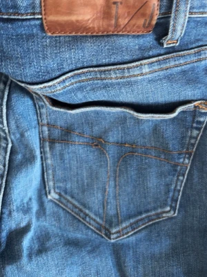 Blå raka jeans från T/J - Säljer ett par klassiska blå jeans från T/J med raka ben och femficksmodell. Jeansen har en snygg mellanblå tvätt, kontrastsömmar och ett brunt läderpatch bak i midjan. Perfekta till en avslappnad stil och passar till det mesta.